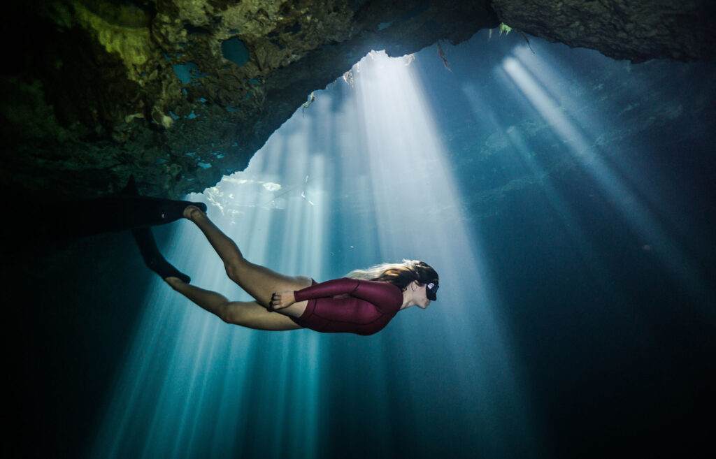 Freediver swimming under a rock ceiling with sunbeams (Cenote Maravilla)