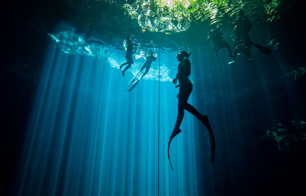 Group of freedivers training at the surface during an AIDA 4 course with Pranamaya in a cenote, with light rays filtering through the water.