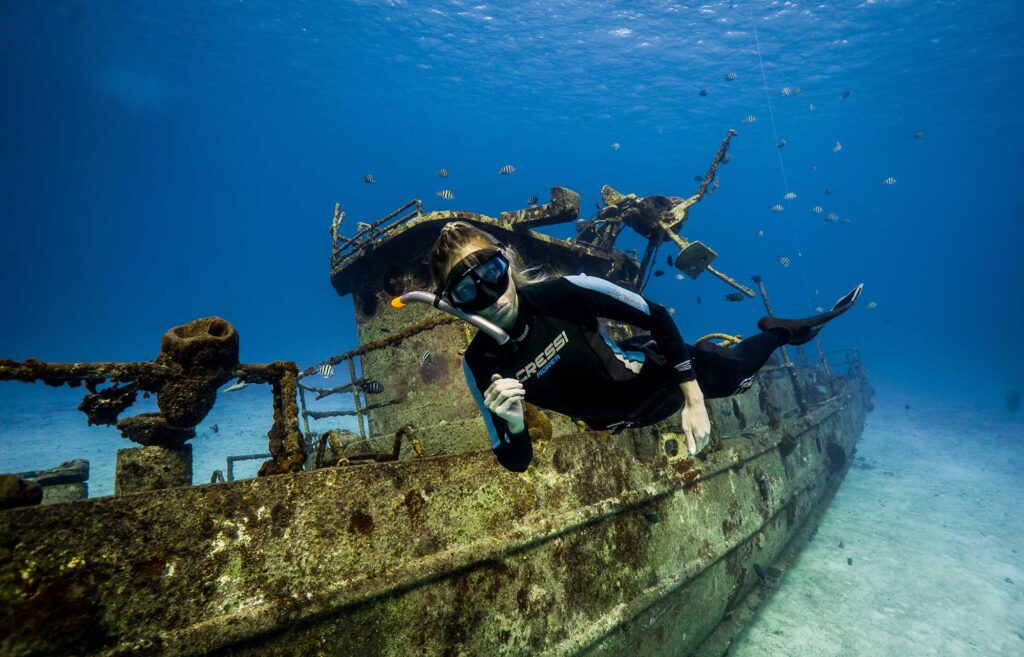 Freediver gliding along a shipwreck surrounded by reef fish in Cozumel waters.