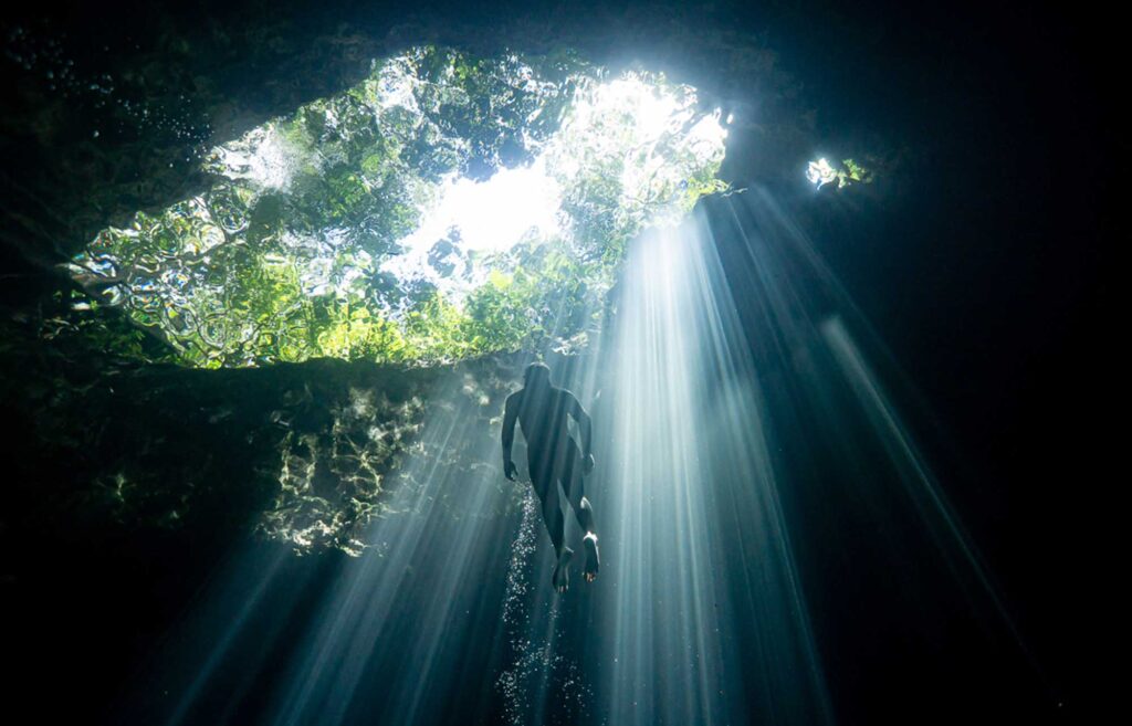Freediver ascending toward a cenote opening as sunlight beams penetrate the water