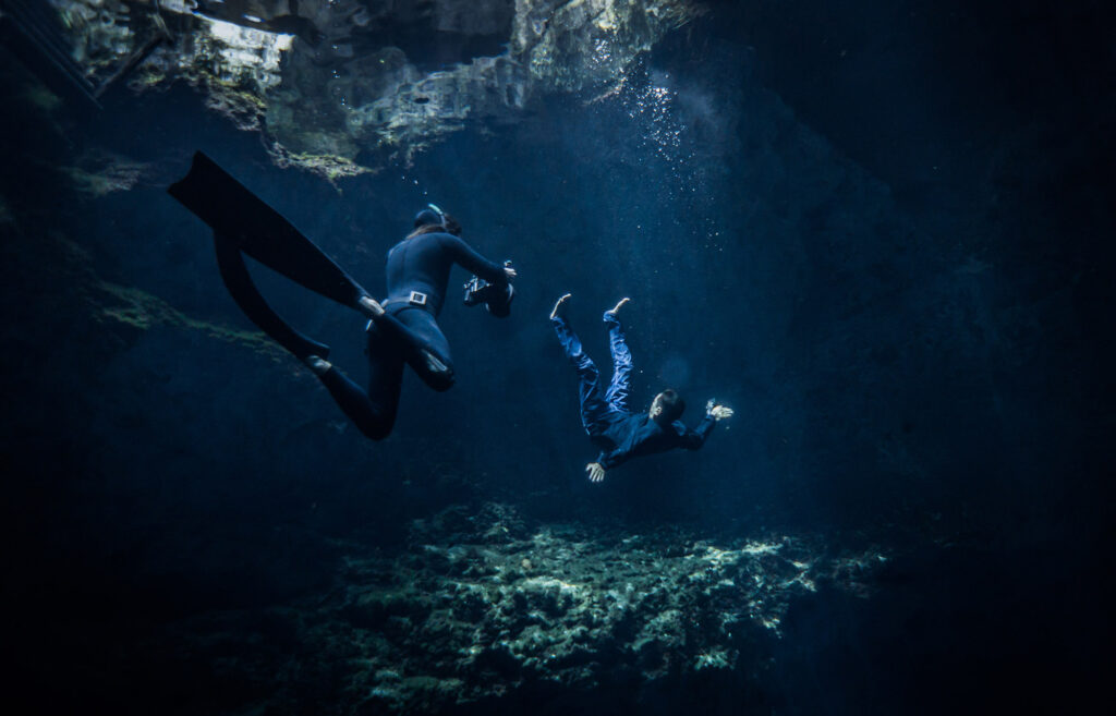 Underwater photographer capturing a freediver descending in a cenote, illuminated by natural light rays